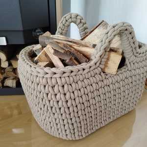 Könnte beinhalten: Ein beigefarbener Häkelkorb mit zwei Griffen, gefüllt mit Brennholz. Der Korb steht auf einem Holzboden.