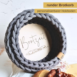 Könnte beinhalten: Ein grauer, runder Brotkorb mit geflochtenem Seildesign. Der Korb hat einen Holzsockel mit dem Wort "Brotzeit" und einer Brotillustration. Der Text "ruder Brotkorb" und "personalisierbarer Holzboden" sind ebenfalls sichtbar.