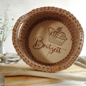 Könnte beinhalten: Ein brauner gehäkelter Brotkorb mit einem Holzboden. Der Boden ist mit dem Text "Brozeit" und einem Bild eines mit Brot gefüllten Korbes graviert.