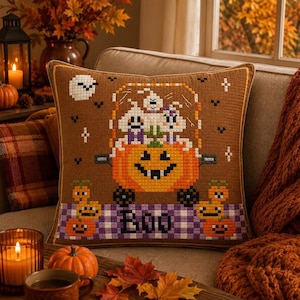 Può includere: Un cuscino quadrato marrone con un disegno a punto croce di tre fantasmi bianchi che cavalcano una grande zucca arancione. Il bordo inferiore presenta un motivo a scacchi viola e bianchi con la parola BOO e piccole zucche arancioni.