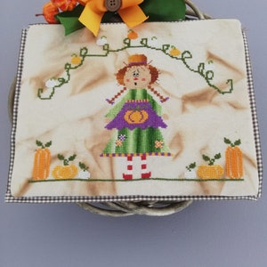 Può includere: Un arazzo da parete a punto croce con una ragazza dei cartoni animati vestita di verde e viola con una zucca in mano. La ragazza è circondata da zucche e viti. L'arazzo è incorniciato da un bordo a scacchi marrone e bianco.