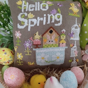 Può includere: Un cuscino grigio con il testo "Hello Spring" ricamato su di esso. Il cuscino presenta una casetta per uccelli, uccelli e fiori. Il cuscino è circondato da uova di Pasqua e un coniglietto soffice.