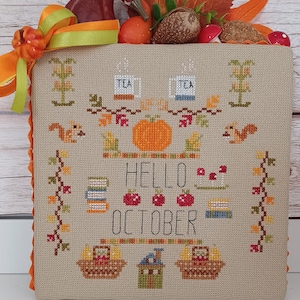 Könnte beinhalten: Kreuzstich-Stickerei mit Herbstthema. Das Design zeigt einen Kürbis, Blätter, Teetassen, Eichhörnchen und den Text "Hello October".
