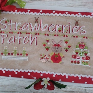 Può includere: Disegno di ricamo a punto croce con un coniglio in un vestito rosso con un cestino di fragole, un campo di fragole e un supporto a più livelli con tazze da tè e fragole. Il disegno è intitolato "Strawberries Patch".