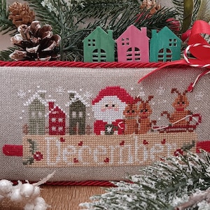 Può includere: Un sampler a punto croce con una scena invernale con Babbo Natale, renne e case. Il sampler è incorniciato con un bordo rosso e ha la parola "Dicembre" ricamata al centro.