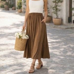 May include: A woman wearing a white square-neck tank top and a brown midi skirt, holding a woven basket filled with white flowers. She is wearing sandals and standing on a cobblestone path, with potted plants and buildings in the background.