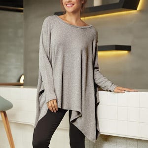 Peut inclure: Un haut tunique gris à manches longues avec un col bateau et un ourlet asymétrique. Le haut est associé à des leggings noirs. Le tissu semble être un tricot doux.