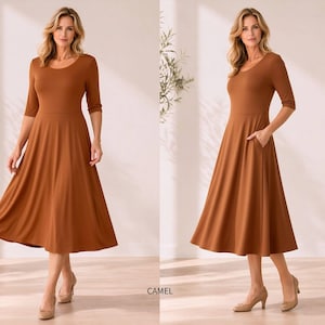 Op de afbeelding: Een camelkleurige jurk met een ronde hals en driekwartmouwen. De jurk heeft een getailleerd lijfje en een uitlopende rok die tot halverwege de kuit valt. De jurk heeft zakken. Het model draagt beige hakken.