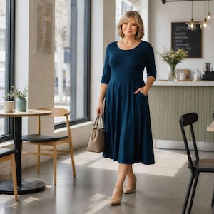 Op de afbeelding: Een vrouw draagt een donkerblauwe, knielange jurk met driekwart mouwen en zakken. Ze houdt een lichtbruine handtas vast en draagt beige hakken. De setting is een caf&eacute; met tafels en stoelen.