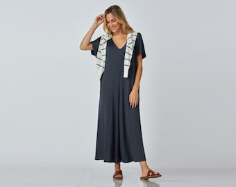 Puede incluir: Mono gris oscuro con cuello en V y mangas cortas. Un suéter color crema con rayas negras está drapeado sobre los hombros. La persona lleva sandalias marrones. El atuendo es simple y elegante.