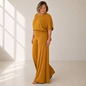 Puede incluir: Un mono de color amarillo mostaza con cuello redondo, mangas cortas tipo dolman y pantalones de pierna ancha. La prenda est&aacute; confeccionada con una tela lisa de color s&oacute;lido que cae holgadamente sobre el cuerpo, creando una silueta continua.