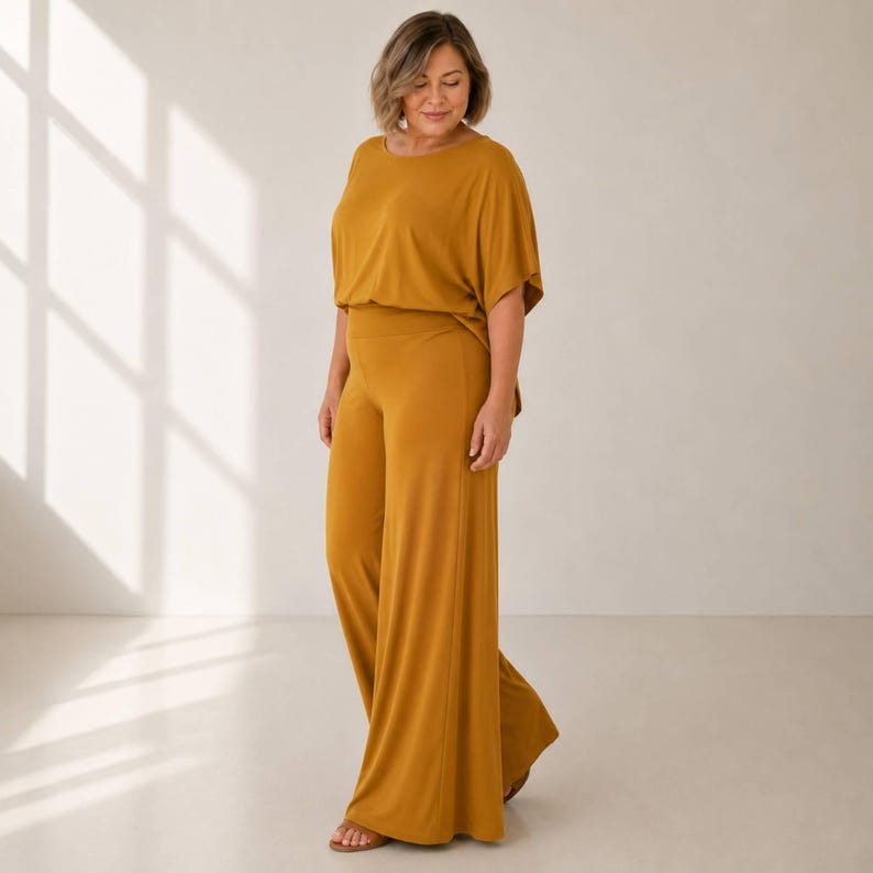 Puede incluir: Un mono de color amarillo mostaza con cuello redondo, mangas cortas tipo dolman y pantalones de pierna ancha. La prenda est&aacute; confeccionada con una tela lisa de color s&oacute;lido que cae holgadamente sobre el cuerpo, creando una silueta continua.