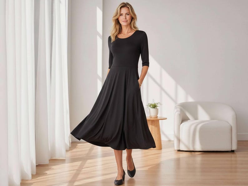 Op de afbeelding: Een zwarte midi-jurk met een ronde hals en driekwart mouwen. De jurk heeft een uitlopende rok en zakken. Het model draagt zwarte ballerina's. De achtergrond omvat witte gordijnen, een witte stoel en een kleine houten tafel.