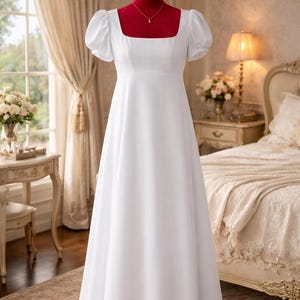 May include: A white, floor-length dress with a square neckline and short, puffed sleeves. The dress is displayed on a red mannequin in a room with a window and a bed. A delicate gold necklace is visible.