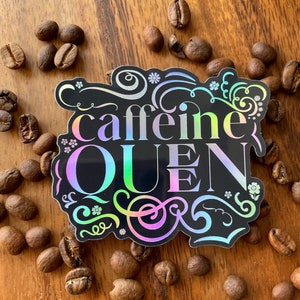 May include: A black and iridescent sticker with the text "caffeine queen" in a decorative font. The sticker is surrounded by coffee beans on a wooden surface.