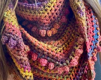 Puede incluir: Un chal de ganchillo hecho a mano y colorido en tonos morado, rosa, naranja y amarillo. El chal presenta un patrón de puntadas texturizadas con bolitas y borlas en los extremos. Este accesorio es ideal para añadir calidez y estilo.