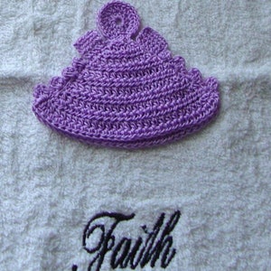 Op de afbeelding: Een paarse gehaakte muts met een bloem erop. De muts ligt op een witte handdoek met het woord "Geloof" erop geborduurd.