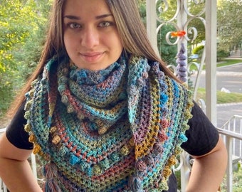 Puede incluir: Un chal de ganchillo multicolor con un diseño texturizado y flecos. El chal está hecho con una variedad de tonos de azul, verde, marrón y naranja.