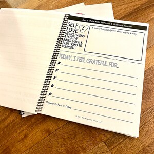 May include: A white journal with a black spiral binding. The cover has the text "Self Love" with a heart symbol and the text "Means having a positive inner voice & being kind to yourself." The journal is titled "Day 1 of the Kid's Self-Love & Gratitude Journal".