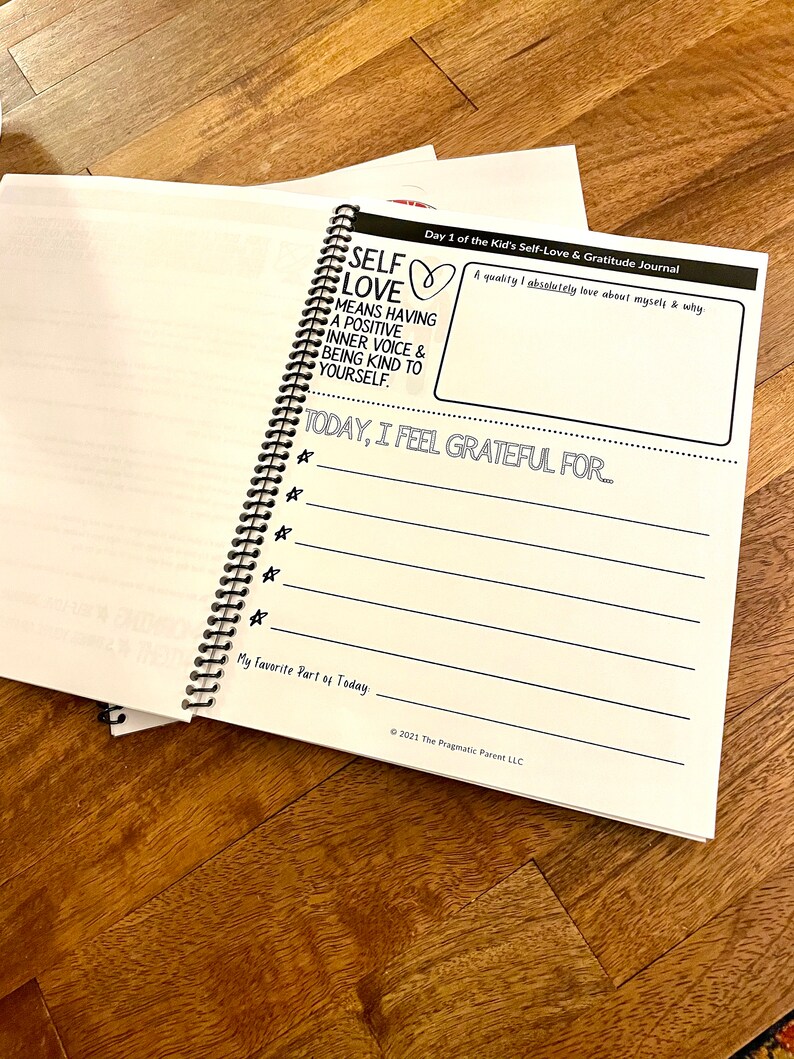 May include: A white journal with a black spiral binding. The cover has the text "Self Love" with a heart symbol and the text "Means having a positive inner voice & being kind to yourself." The journal is titled "Day 1 of the Kid's Self-Love & Gratitude Journal".