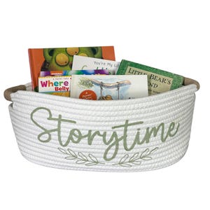 Puede incluir: Una cesta de cuerda blanca llena de libros infantiles. La cesta tiene bordado en verde claro la palabra "Storytime" con detalles de hojas. Los libros tienen cubiertas coloridas, incluyendo "Where's Belly". La cesta tiene un asa de cuerda marr&oacute;n.