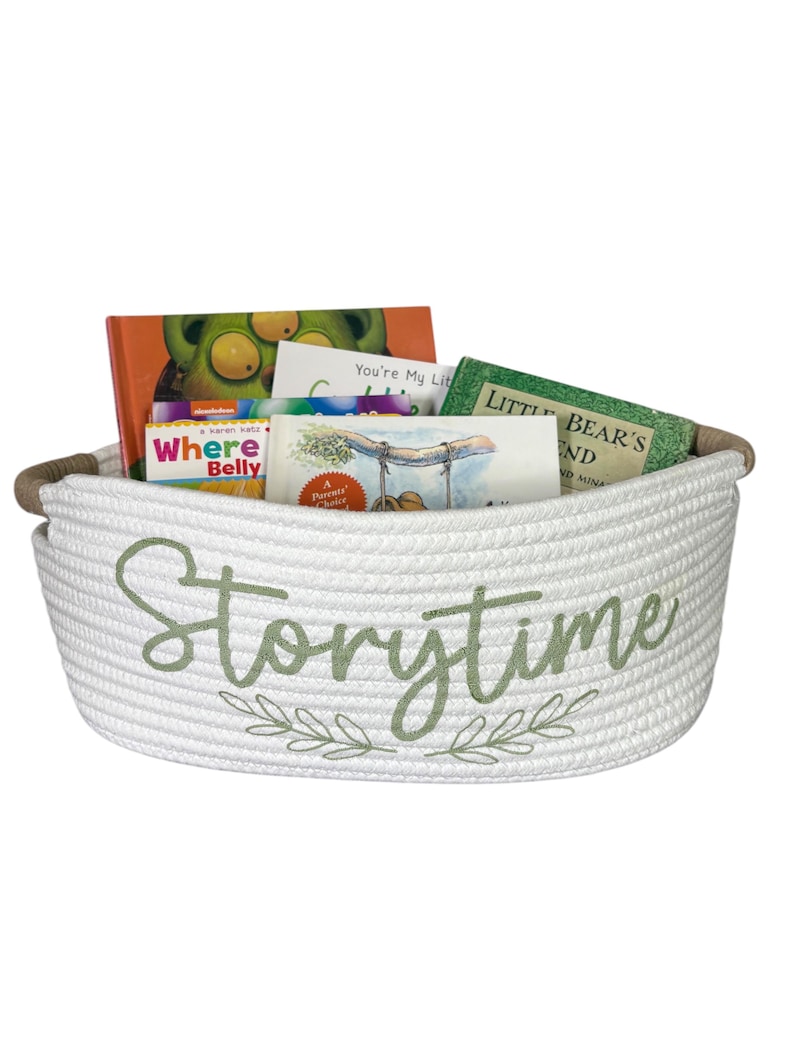 Puede incluir: Una cesta de cuerda blanca llena de libros infantiles. La cesta tiene bordado en verde claro la palabra "Storytime" con detalles de hojas. Los libros tienen cubiertas coloridas, incluyendo "Where's Belly". La cesta tiene un asa de cuerda marr&oacute;n.