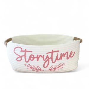 Puede incluir: Una cesta de almacenamiento blanca tejida con la palabra "Storytime" en escritura rosa en la parte delantera. La cesta tiene forma rectangular con esquinas redondeadas y asas de cuerda marr&oacute;n. Un dise&ntilde;o floral rosa est&aacute; debajo de la palabra "Storytime".