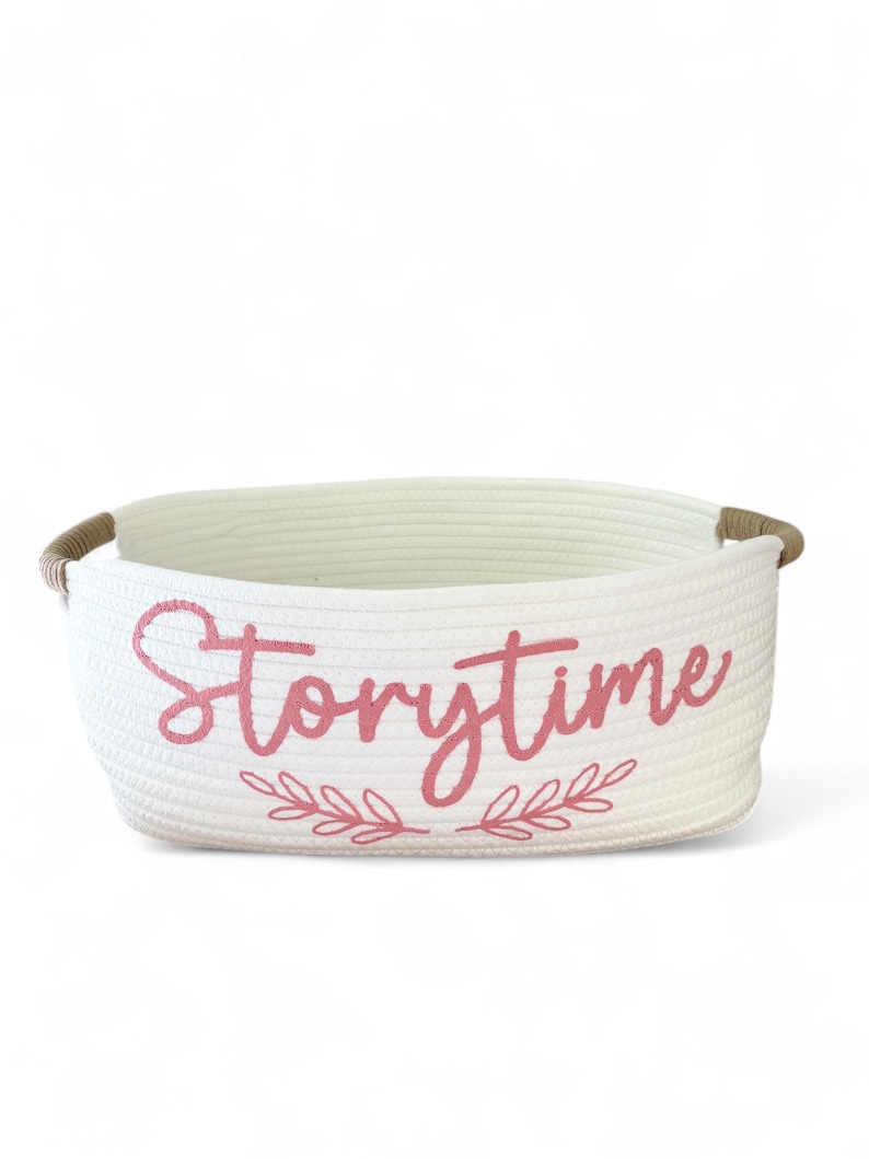 Puede incluir: Una cesta de almacenamiento blanca tejida con la palabra "Storytime" en escritura rosa en la parte delantera. La cesta tiene forma rectangular con esquinas redondeadas y asas de cuerda marr&oacute;n. Un dise&ntilde;o floral rosa est&aacute; debajo de la palabra "Storytime".