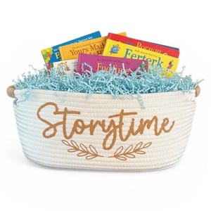 May include: A white rope basket filled with children's books and blue paper filler. The basket has wooden handles and the word "Storytime" in brown script with a leaf design.