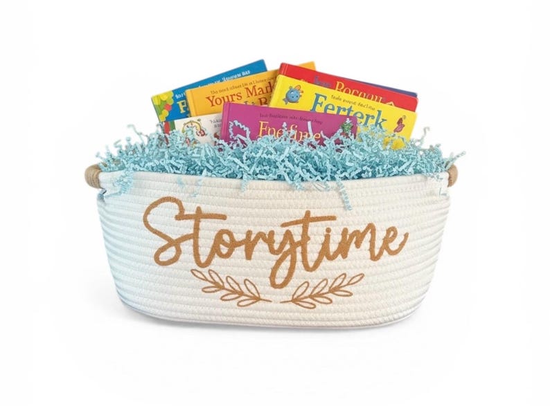 Puede incluir: Una cesta de cuerda blanca llena de libros infantiles y relleno de papel azul. La cesta tiene asas de madera y la palabra "Storytime" en escritura marr&oacute;n con un dise&ntilde;o de hojas.