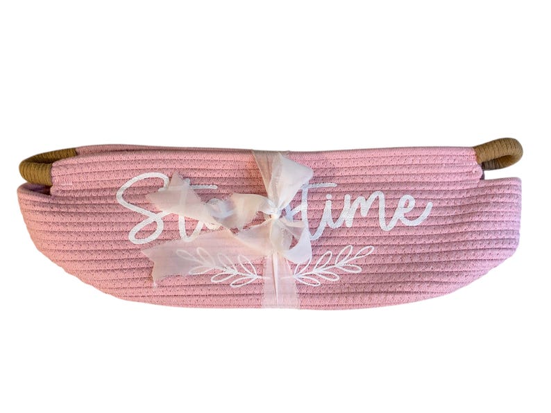 Puede incluir: Una cesta rosa tejida con las palabras "Sweet Time" en escritura blanca. La cesta tiene una cinta blanca atada y asas marrones.