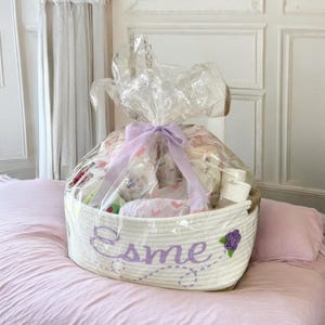 Peut inclure: Un panier en corde blanc rempli d'articles pour b&eacute;b&eacute;s, envelopp&eacute; de cellophane transparent et nou&eacute; avec un ruban lavande. Le panier porte le nom "Esme" brod&eacute; en fil lavande, avec une fleur violette.