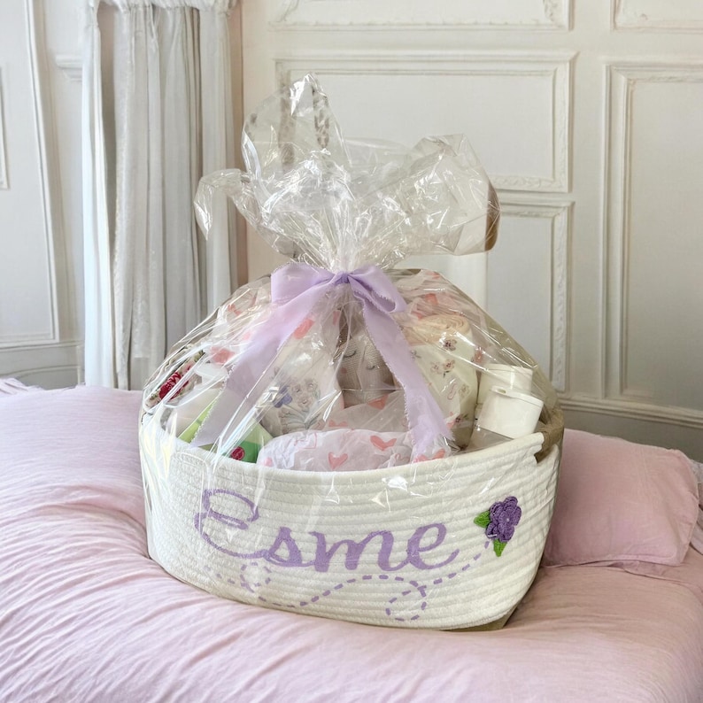 Peut inclure: Un panier en corde blanc rempli d'articles pour b&eacute;b&eacute;s, envelopp&eacute; de cellophane transparent et nou&eacute; avec un ruban lavande. Le panier porte le nom "Esme" brod&eacute; en fil lavande, avec une fleur violette.