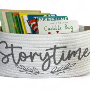 Puede incluir: Una cesta de cuerda blanca llena de libros infantiles coloridos. La cesta tiene la palabra "Storytime" en escritura gris con un dise&ntilde;o de hojas. Una tarjeta dice "You're My Little Cuddle Bug."