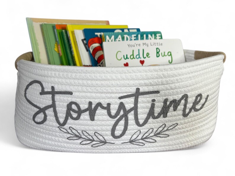 Puede incluir: Una cesta de cuerda blanca llena de libros infantiles coloridos. La cesta tiene la palabra "Storytime" en escritura gris con un dise&ntilde;o de hojas. Una tarjeta dice "You're My Little Cuddle Bug."