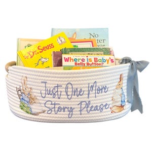 Peut inclure: Un panier en corde blanc rempli de livres pour enfants, dont un livre du Dr. Seuss. Le panier pr&eacute;sente des illustrations de Pierre Lapin et le texte "Just One More Story Please" en &eacute;criture bleue. Un ruban bleu est nou&eacute; sur le c&ocirc;t&eacute;.