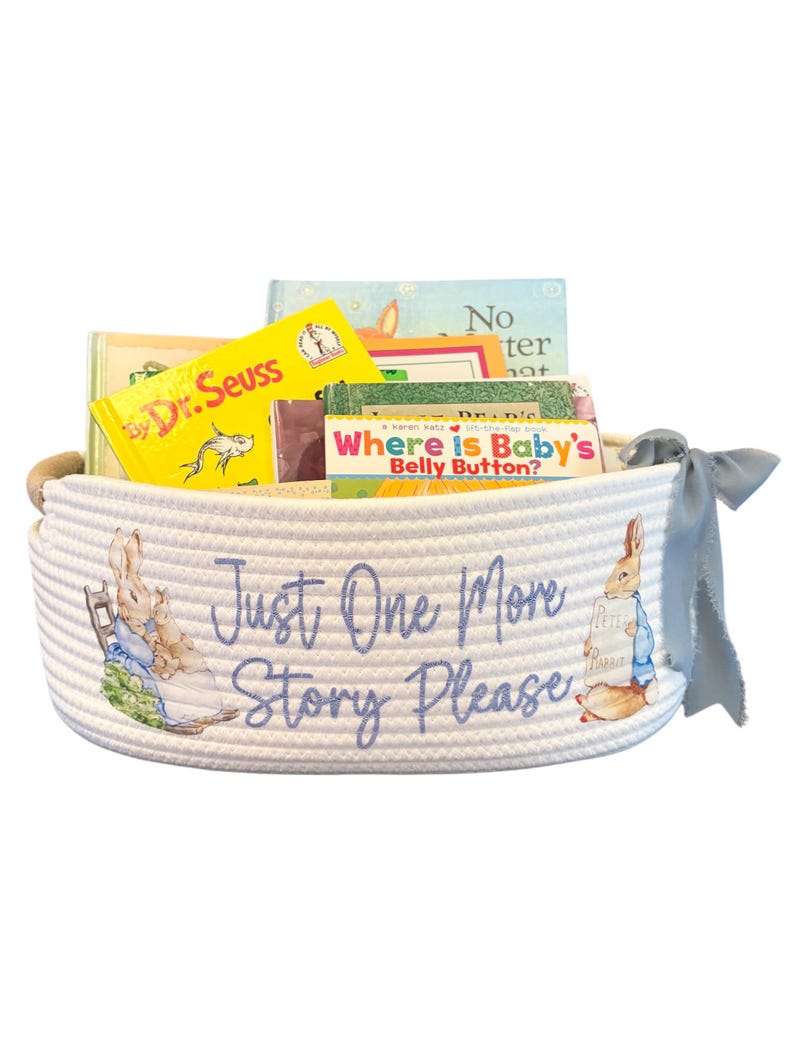 Peut inclure: Un panier en corde blanc rempli de livres pour enfants, dont un livre du Dr. Seuss. Le panier pr&eacute;sente des illustrations de Pierre Lapin et le texte "Just One More Story Please" en &eacute;criture bleue. Un ruban bleu est nou&eacute; sur le c&ocirc;t&eacute;.