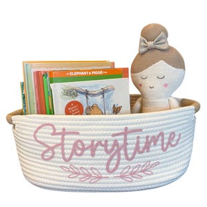 May include: A white rope basket with the word "Storytime" in pink script, holding colorful children's books and a knitted doll with a bow. The basket has a pink floral design below the text, perfect for storing books and toys.