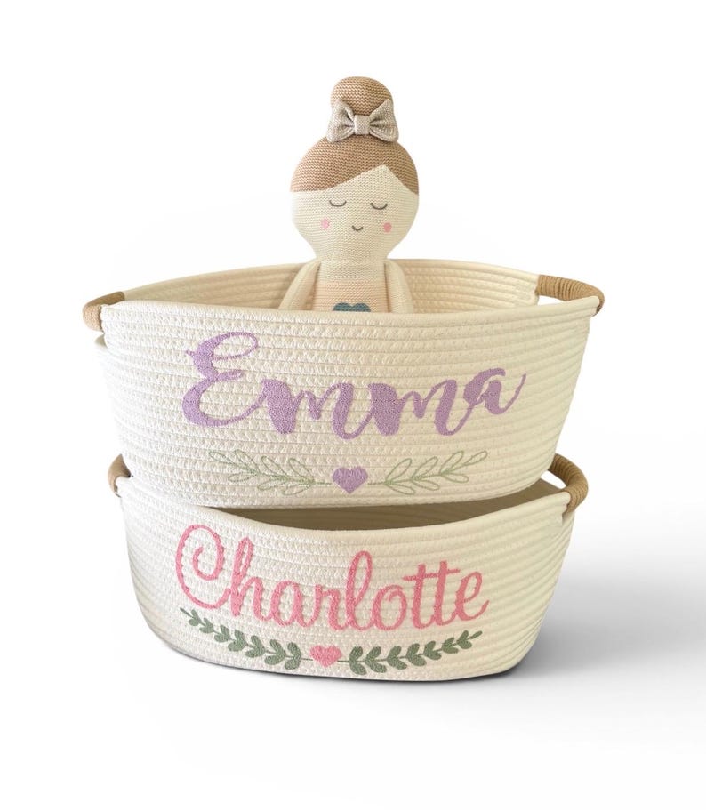 Peut inclure: Deux paniers en corde cr&egrave;me superpos&eacute;s, brod&eacute;s des noms "Emma" et "Charlotte". Une poup&eacute;e avec un chignon et un n&oelig;ud beiges est assise dans le panier sup&eacute;rieur. Les paniers ont des poign&eacute;es marron.