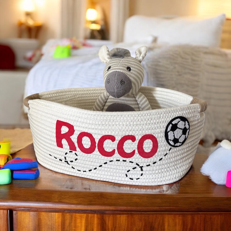 Peut inclure: Un panier en corde cr&egrave;me avec le nom "Rocco" en rouge et un ballon de football. &Agrave; l'int&eacute;rieur, un jouet z&egrave;bre ray&eacute;. Le panier est pos&eacute; sur une surface en bois. Id&eacute;al pour le rangement.