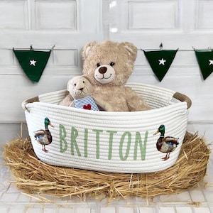 Peut inclure: Un panier en corde blanc avec le nom "Britton" en lettres vertes, d&eacute;cor&eacute; d'illustrations de canards. Le panier contient un ours en peluche et un petit animal en peluche. Le panier repose sur un lit de foin.