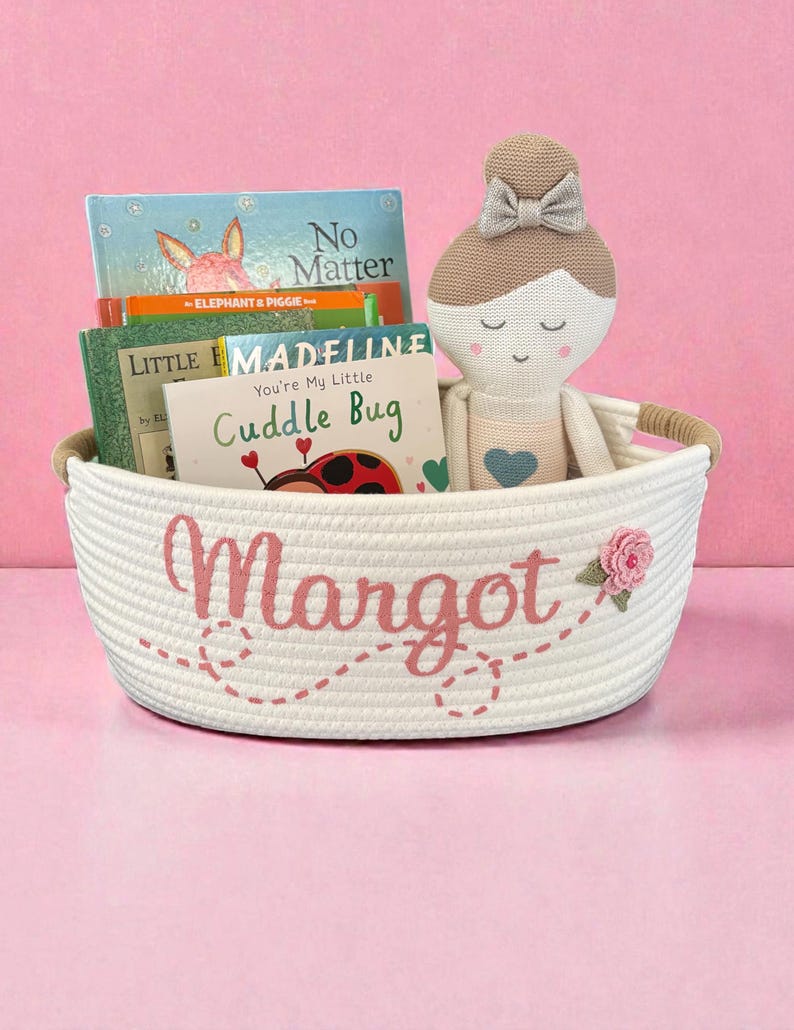Peut inclure: Un panier en corde blanc brod&eacute; du nom "Margot" en rose, contenant des livres pour enfants et une poup&eacute;e. La poup&eacute;e a une t&ecirc;te beige, un n&oelig;ud et un c&oelig;ur bleu. Une rose rose est brod&eacute;e sur le panier.