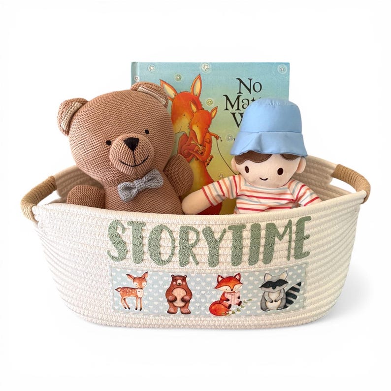 Peut inclure: Un panier en osier cr&egrave;me, &eacute;tiquet&eacute; "Storytime", contient un ours en peluche marron, une poup&eacute;e avec un chapeau bleu et une chemise ray&eacute;e, et un livre pour enfants. Le panier pr&eacute;sente un panneau avec des illustrations d'animaux de la for&ecirc;t.