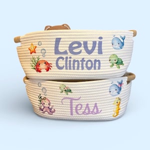 Peut inclure: Deux paniers en corde blanche empil&eacute;s. Le panier sup&eacute;rieur porte le nom "Levi Clinton" en violet, avec des illustrations de cr&eacute;atures marines. Le panier inf&eacute;rieur porte le nom "Tess" en violet, avec des illustrations de cr&eacute;atures marines.