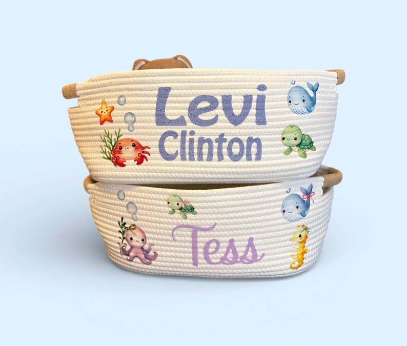 Peut inclure: Deux paniers en corde blanche empil&eacute;s. Le panier sup&eacute;rieur porte le nom "Levi Clinton" en violet, avec des illustrations de cr&eacute;atures marines. Le panier inf&eacute;rieur porte le nom "Tess" en violet, avec des illustrations de cr&eacute;atures marines.