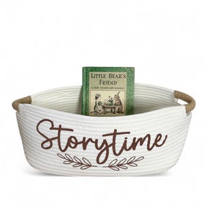 Puede incluir: Una cesta de cuerda blanca con asas marrones y la palabra "Storytime" en escritura marr&oacute;n. Un libro verde titulado "Little Bear's Friend" est&aacute; dentro de la cesta. La cesta tiene un dise&ntilde;o de hojas decorativas.