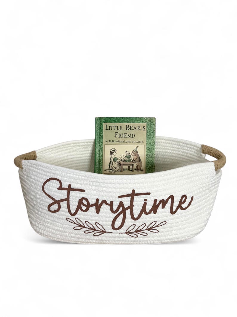Puede incluir: Una cesta de cuerda blanca con asas marrones y la palabra "Storytime" en escritura marr&oacute;n. Un libro verde titulado "Little Bear's Friend" est&aacute; dentro de la cesta. La cesta tiene un dise&ntilde;o de hojas decorativas.