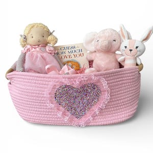 May include: A pink woven basket filled with a doll, a plush elephant, a plush bunny, and a book titled "Guess How Much I Love You." The basket has a heart-shaped decoration with glitter and lace.