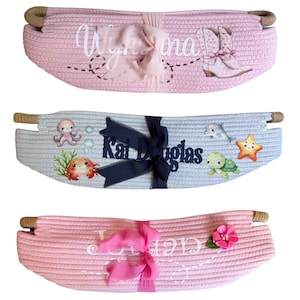 Peut inclure: Trois paniers en corde tress&eacute;e roses et bleus, chacun avec des &eacute;l&eacute;ments d&eacute;coratifs. Un panier rose porte un nom et des bottes de cowboy, un autre a un poulpe et un crabe, et le troisi&egrave;me a un nom et une fleur. Chaque panier a des poign&eacute;es en corde.