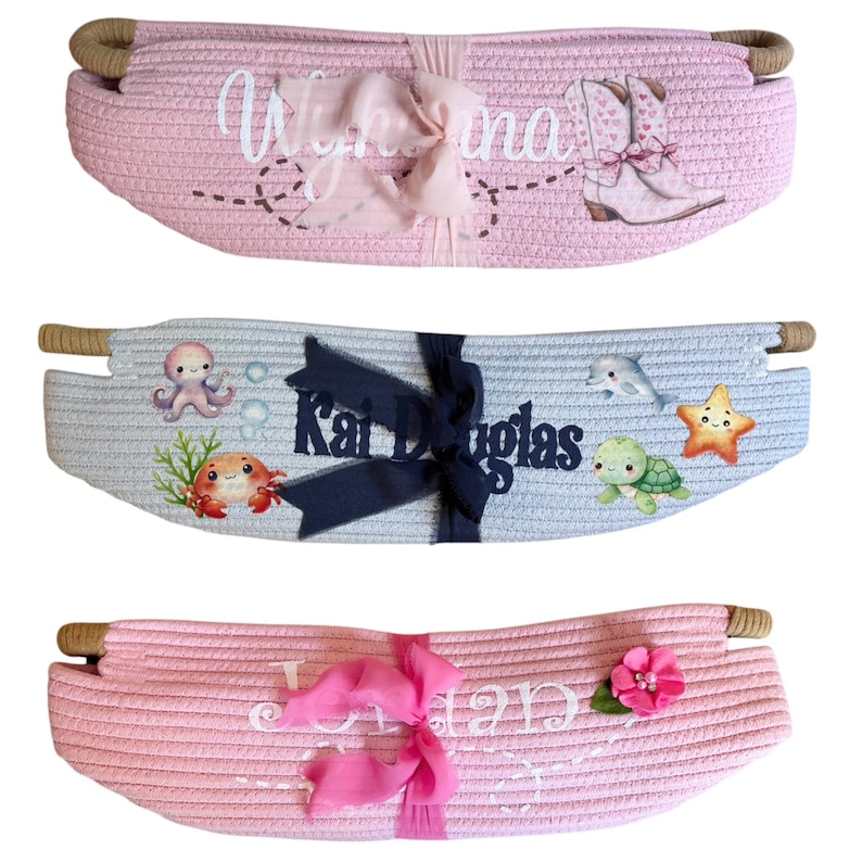 Peut inclure: Trois paniers en corde tress&eacute;e roses et bleus, chacun avec des &eacute;l&eacute;ments d&eacute;coratifs. Un panier rose porte un nom et des bottes de cowboy, un autre a un poulpe et un crabe, et le troisi&egrave;me a un nom et une fleur. Chaque panier a des poign&eacute;es en corde.